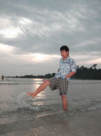 Man splashing water with leg while standing on shore at beach during sunset against cloudy sky