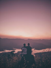 Rear view of friends sitting at cliff against sky during sunset