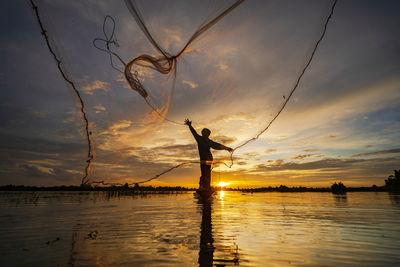 Silhouette man throwing fishing net at river against sky during sunset