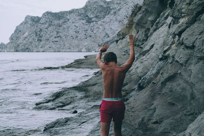 Rear view of shirtless teenager walking by cliff on shore