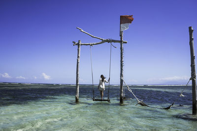 Rear view of woman standing on swing over sea against sky