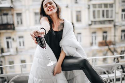 Cheerful woman having red wine while wrapped in blanket at balcony