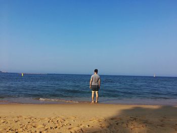 Rear view of mature man standing on shore at beach against clear blue sky