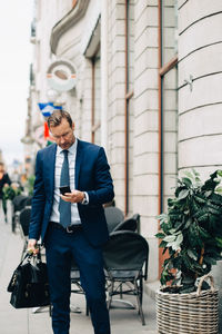 Mature businessman using smart phone while walking with bag on sidewalk in city