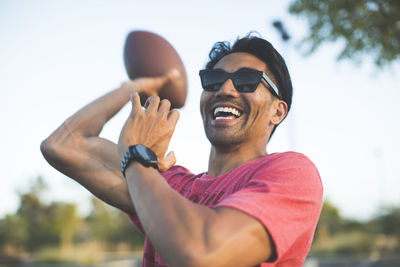 Cheerful man in sunglasses playing with american football against sky