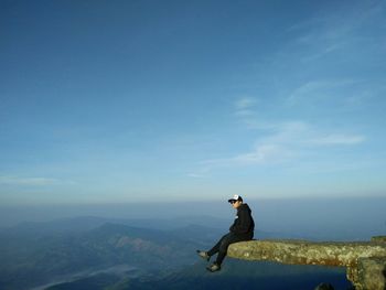 Side view of man sitting on cliff against sky