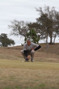 Mature man crouching on grassy field against cloudy sky