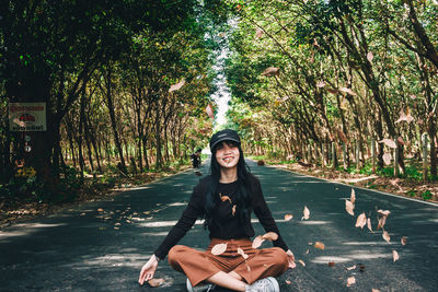 Portrait of smiling young woman throwing leaves while sitting on road amidst trees in forest