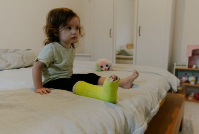A thoughtful girl with a cast on her leg is sitting on the bed.