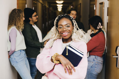 Portrait of smiling young woman with diary while multiracial friends in background at corridor in college dorm