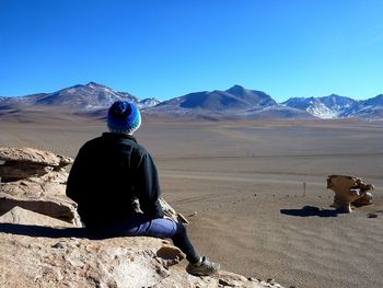 Rear view full length of man sitting on rock at desert against clear blue sky