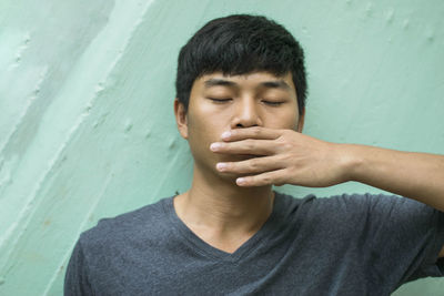 Close-up of young man with hand covering mouth standing against wall