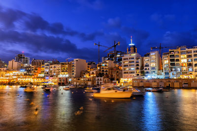 Sailboats moored on sea by illuminated buildings against sky at dusk