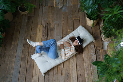 Joyful young female wearing virtual reality glasses resting on cozy soft mattress in home greenhouse