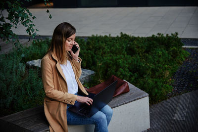 Woman sitting on bench in city street, using laptop and smartphone