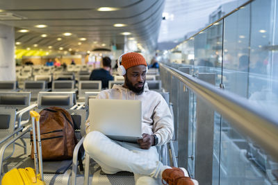 Young hipster black man sitting in airport terminal using laptop wear headphones and listening music