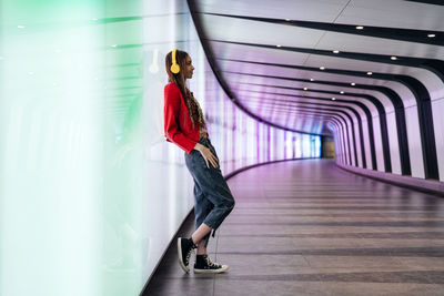 Thoughtful woman with headphones leaning on illuminated wall in underground tunnel
