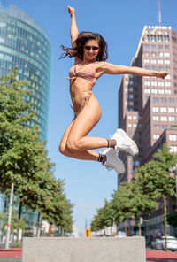 Young woman wearing bikini while jumping against buildings in city during sunny day