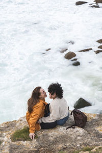 Happy young female friends sitting on rock by sea during weekend