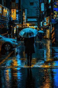 Rear view of woman with umbrella walking on illuminated street during rainy season