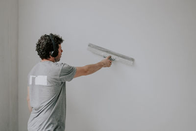 Young male plasterer listens to music through headphones and holds a large spatula on the wall