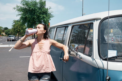 Young woman drinking fesh water from water bottle near a camper van in urban parking. traveling. 