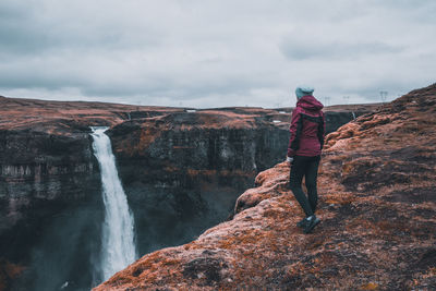 Rear view of woman looking at waterfall while standing on rock against sky