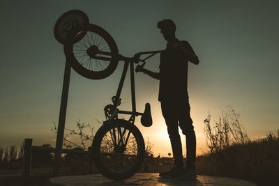 Silhouette man standing by bicycle against sky during sunset