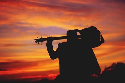 Rear view of silhouette man holding guitar against sky during sunset