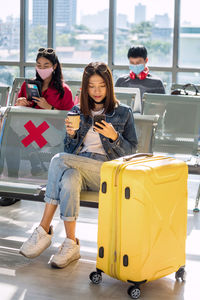Asian young tourist girl using phone and hold hot coffee glass at departure waiting seats at airport