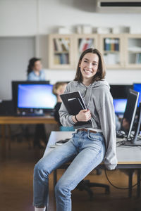 Cheerful high school female student sitting with books on desk in classroom