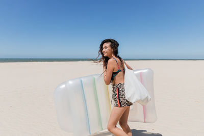 Side view of positive female in summer outfit and with inflatable mattress walking on sandy coast on sunny day during vacation