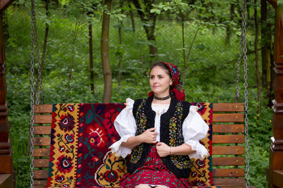 Thoughtful young woman in romanian clothing sitting on swing at yard