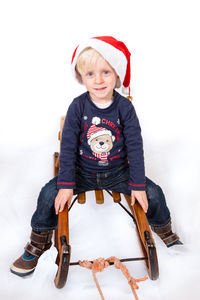 Full length portrait of cute boy wearing santa hat while sitting on sledge against white background