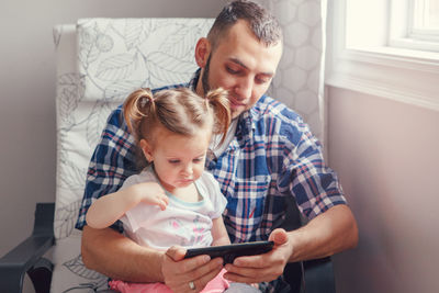 Man with daughter using phone while sitting on chair at home