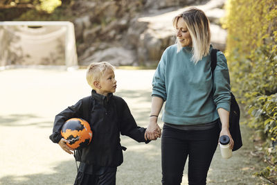 Mother holding hand of son carrying soccer ball while walking in ground