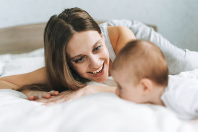 Young mother having fun with cute baby girl on bed with white linen, natural tones, love emotion