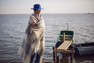 Woman in jeans and a sweater and a hat stands on a pier with boats by the lake in autumn