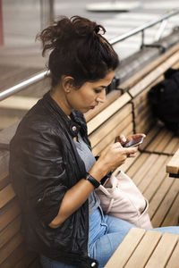 Woman using phone while sitting on wooden bench of sidewalk cafe in city