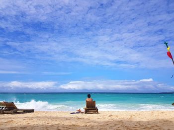 Rear view of woman sitting on deck chair at beach against sky