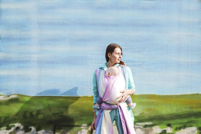 Mother carrying baby wrapped in blanket looking away while standing against wall