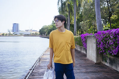 Young man looking away while standing on boardwalk by lake