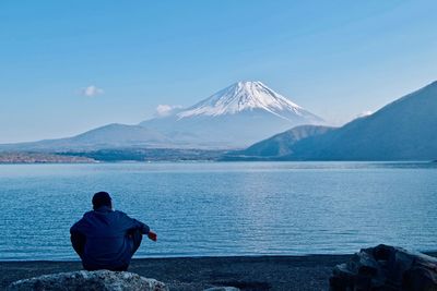 Rear view of man sitting by lake against mountain range