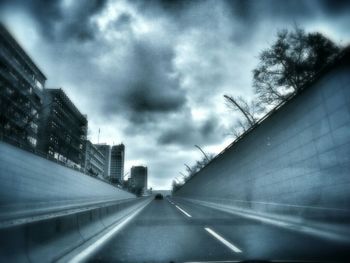 Road amidst buildings in city against cloudy sky