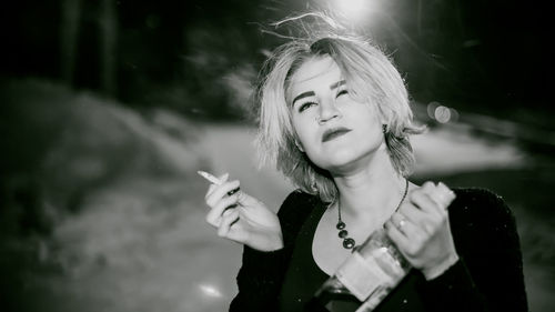 Close-up of young woman smoking cigarette while holding alcohol bottle at night