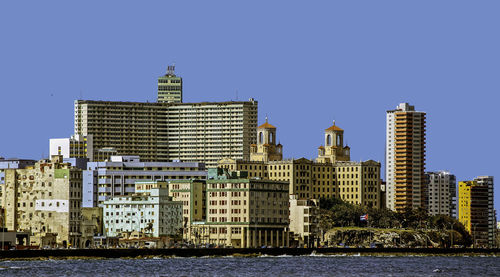 Modern buildings by river against clear blue sky in city during sunny day