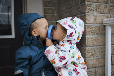 Playful siblings wearing raincoats while kissing through magnifying glass in porch