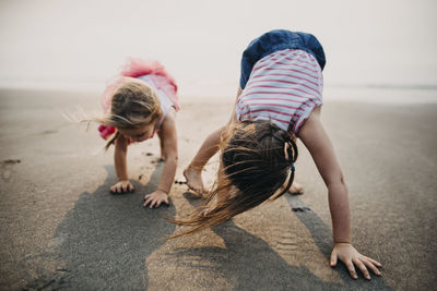 Playful sisters bending on sand at beach against sky