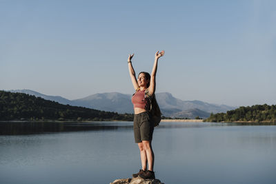 Smiling female hiker with arms raised standing on rock at lake