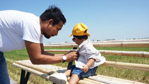 Father dressing son sitting on bench against clear blue sky